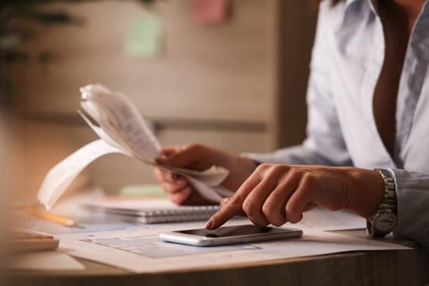 Close up of businesswoman using smart phone while calculating her bills in the office. Close up of businesswoman going though finances and using mobile phone as calculator. Professional Invoices stock pictures, royalty-free photos & images